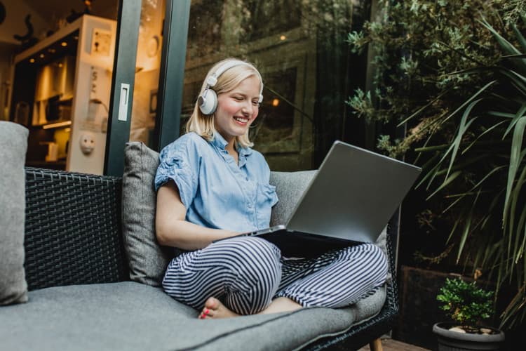 Person sitzt mit Kopfhörern und Laptop auf einer Terrasse zwischen Pflanzen.