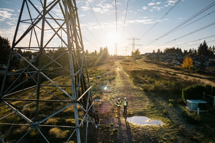 Zwei Personen in Warnkleidung stehen unter Hochspannungsleitungen neben einem Strommast