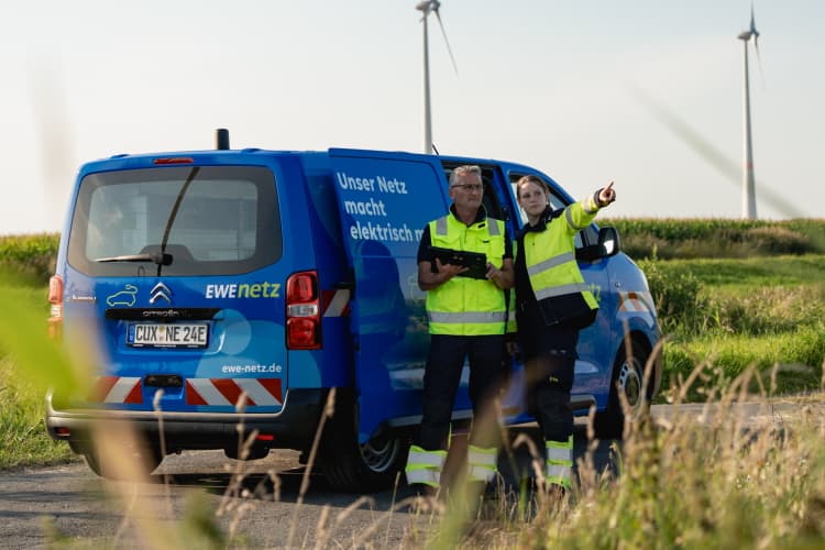 Zwei Mitarbeitende von EWE NETZ vor einem Servicefahrzeug und Windkraftanlagen im Einsatz vor Ort