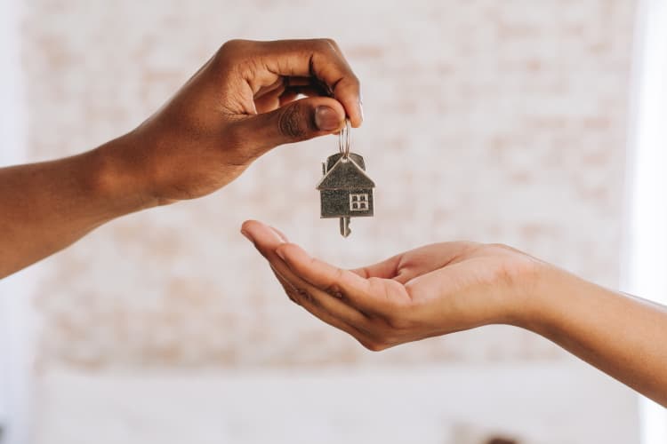 Schlüsselübergabe mit Hausanhänger zwischen zwei Personen als Symbol für Eigentümerwechsel oder Immobilienübergabe.