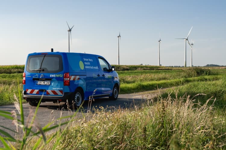 Ein blauer Servicewagen fährt auf einem schmalen Weg durch eine grüne Landschaft. Im Hintergrund sind mehrere Windenergieanlagen auf Feldern zu sehen.