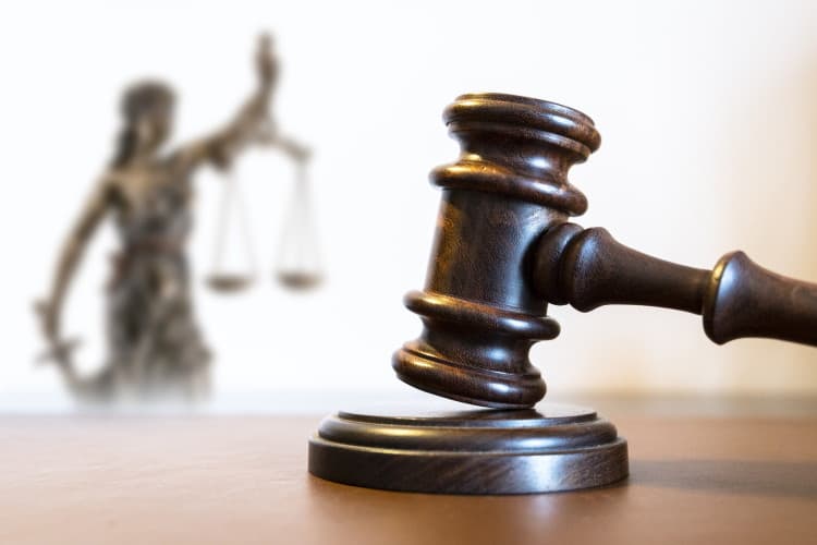 Richterhammer steht auf einer Oberfläche im Vordergrund. Im Hintergrund ist eine Justitia-Figur mit Waage