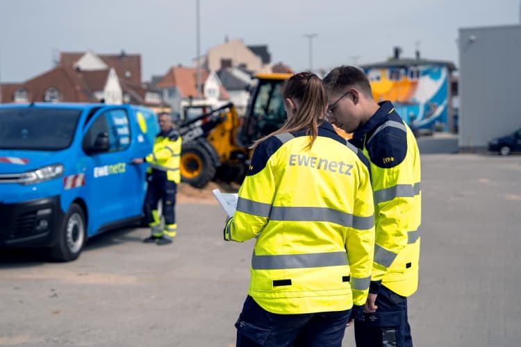 Zwei Personen in Warnschutzkleidung stehen auf einer Baustelle und betrachten gemeinsam Unterlagen